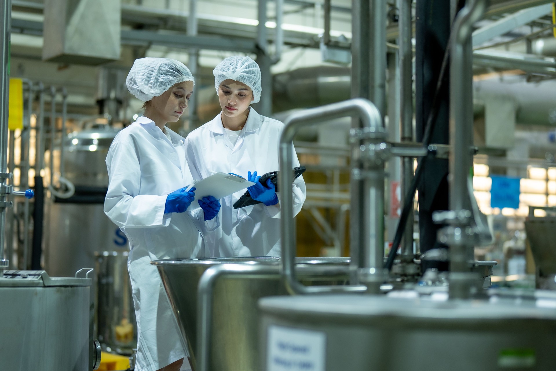 Two female scientists wearing protective equipment and use a tablet and clipboard to verify the correctness of the ingredients in the beverage mixing tank.